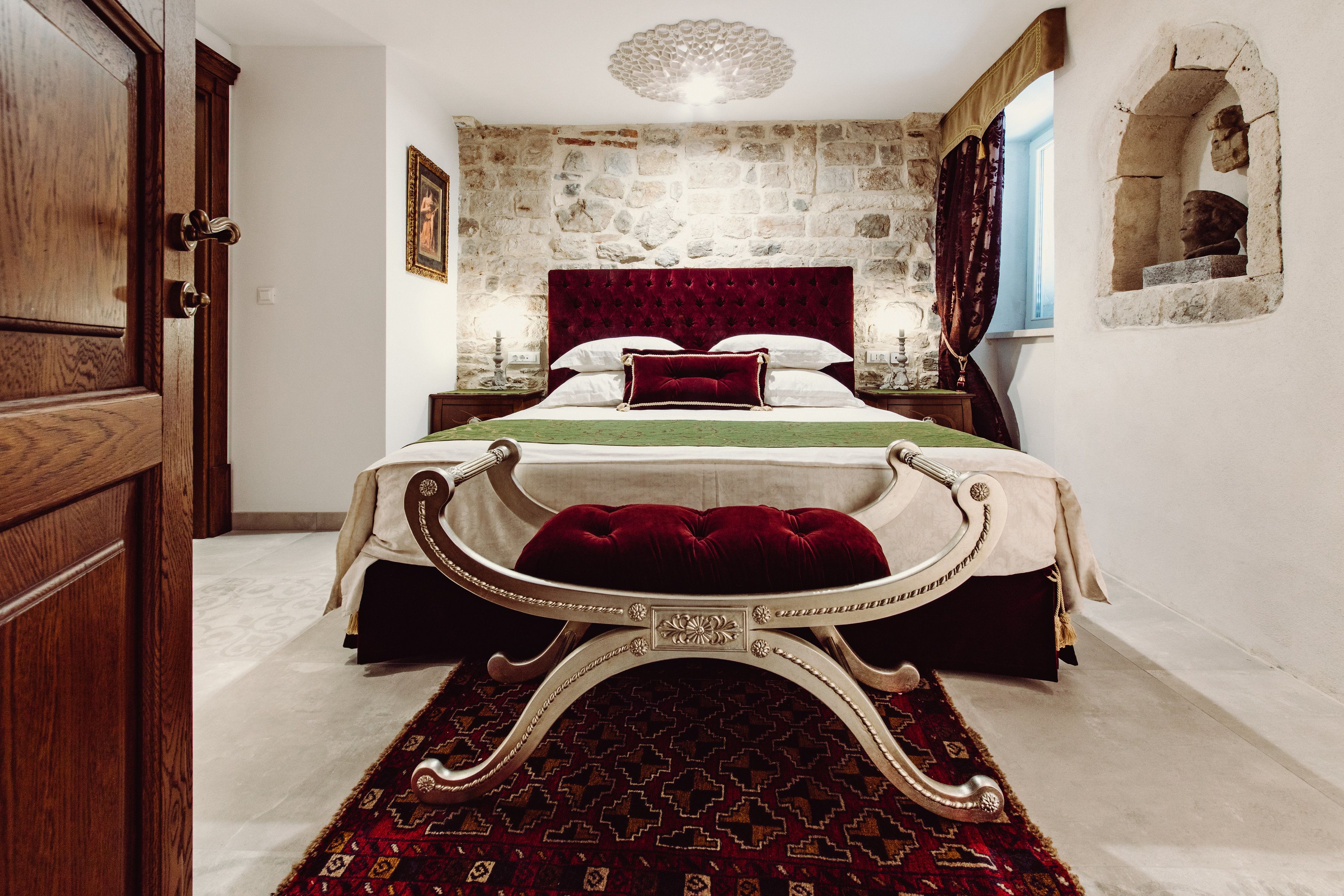 Une chambre avec un mur en pierre, un grand lit avec une tête de lit rouge, un plaid vert et un tapis à motifs.