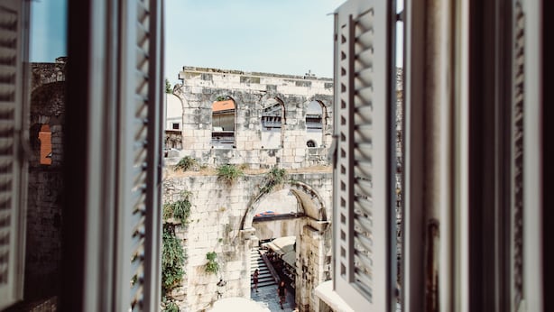 Una ventana abierta con vistas a un edificio de piedra con arcos y una escalera que lleva a una puerta.