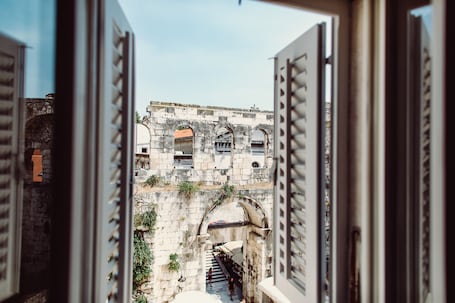 An open window with a view of a stone building with arches and a staircase leading to a door.