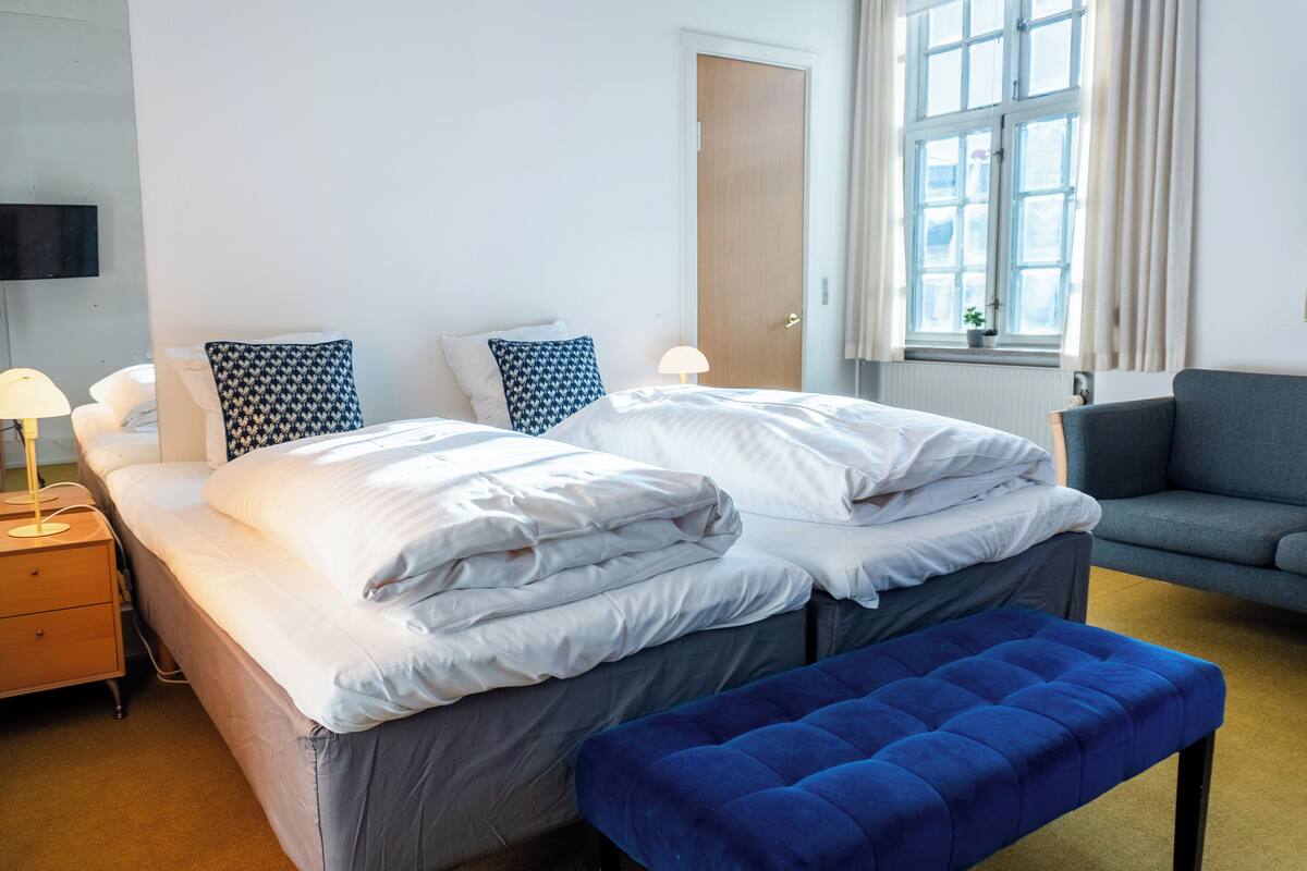 A hotel room with a bed, a bench, a sofa, a TV, and a window with curtains.