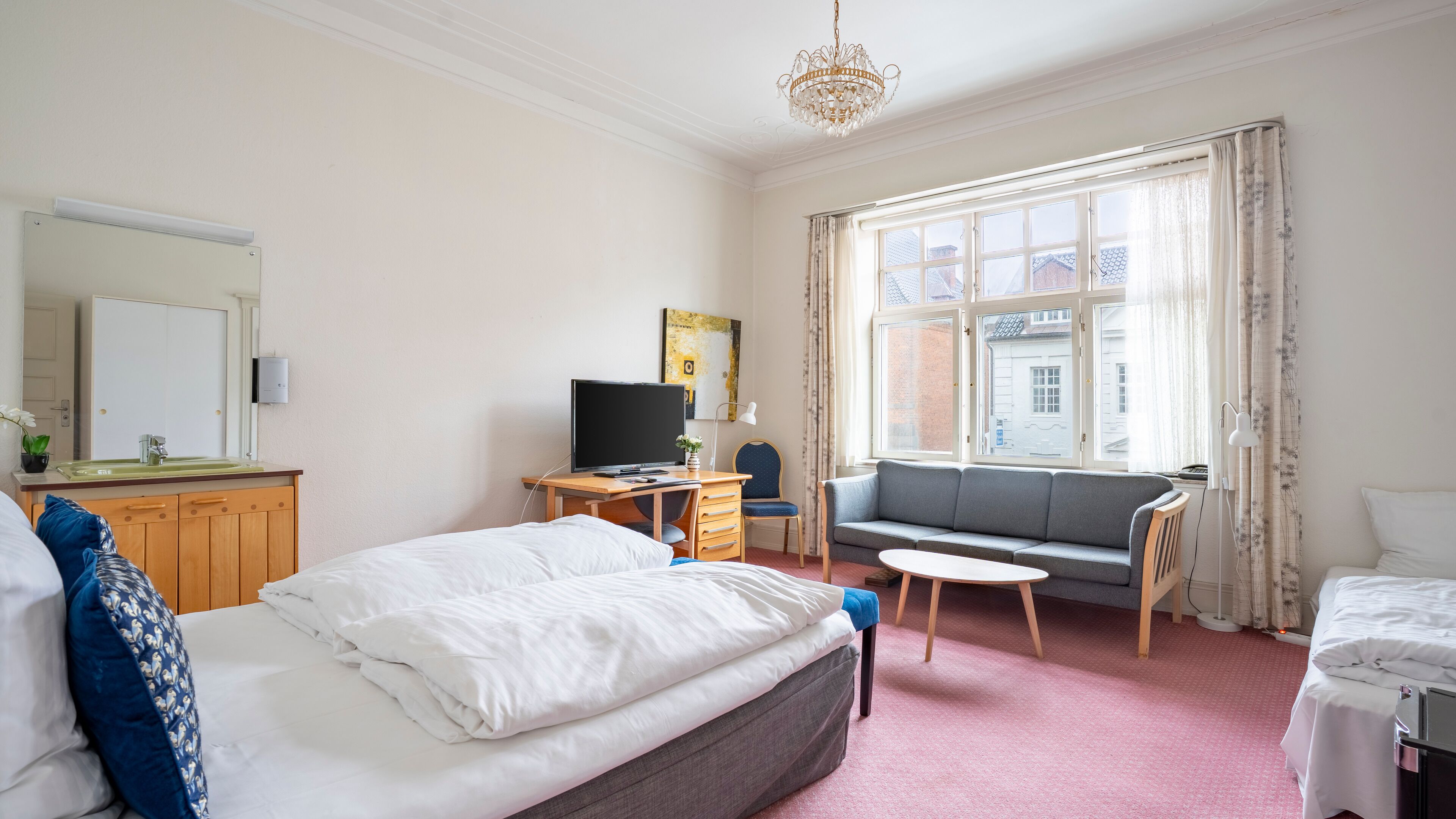 A hotel room with a bed, a desk with a computer, a sofa, and a large window with curtains.