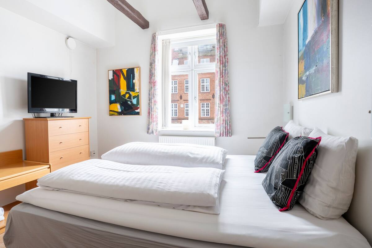 A hotel room with a bed, a wooden desk, a television, a window with curtains, and artwork on the walls.