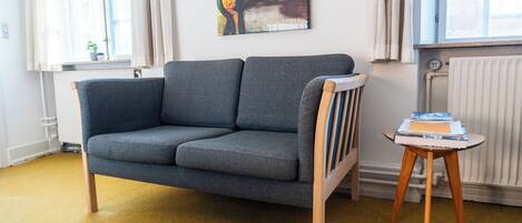 Una sala moderna con un sofá gris, una pequeña mesa auxiliar de madera con libros, un cuadro abstracto y colorido en la pared, y una ventana con cortinas.