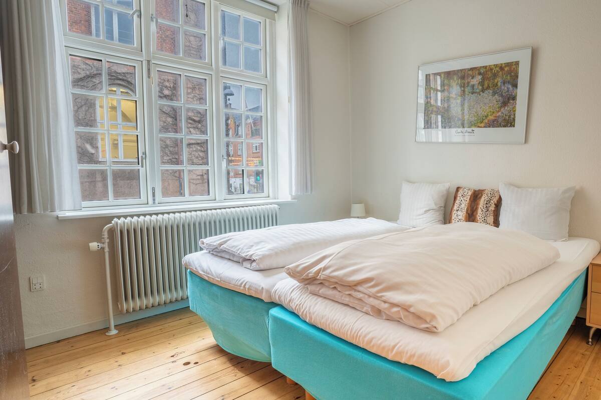 A bedroom with a double bed, a nightstand, a radiator, and a window with a view of buildings.