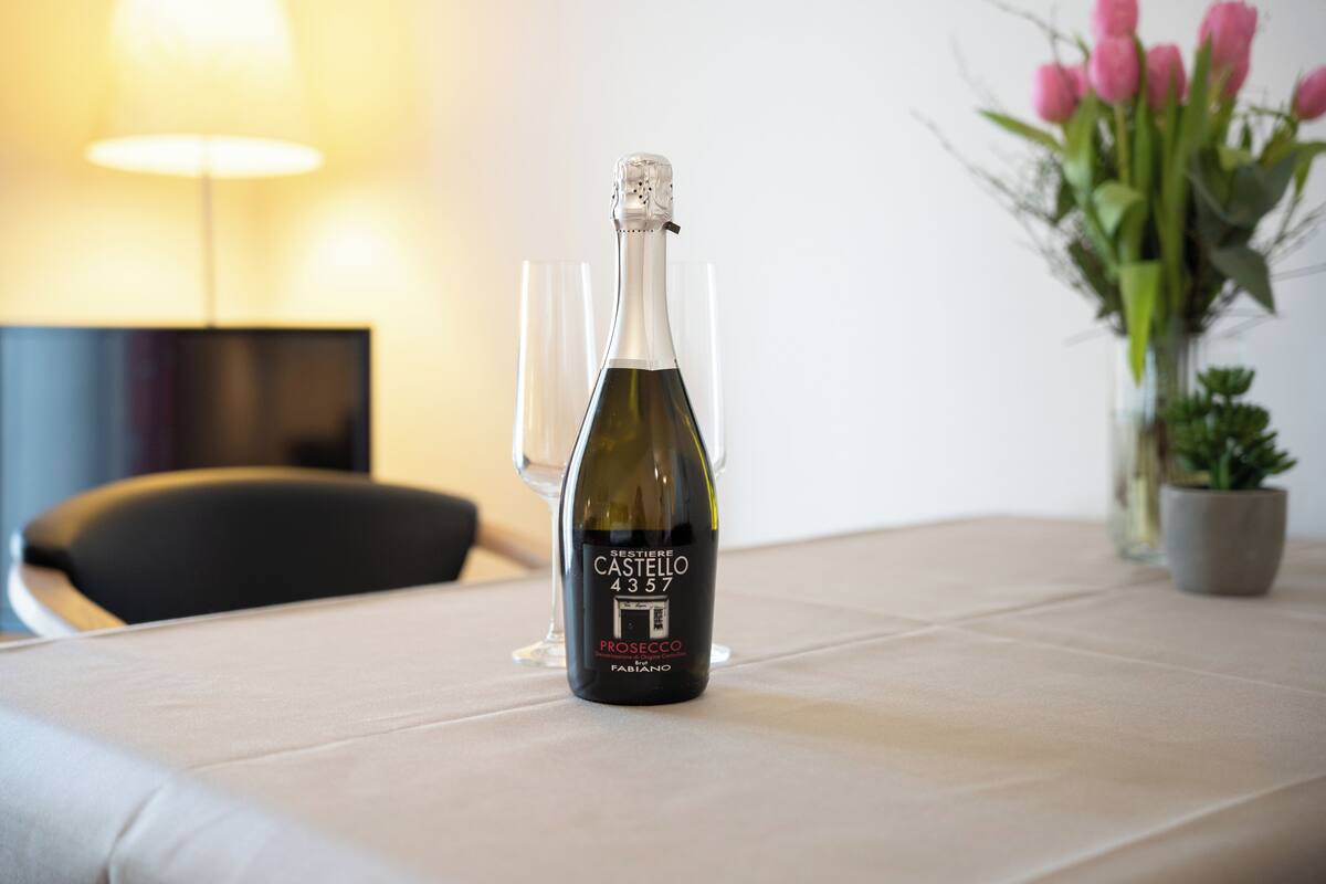 A bottle of wine on a table with a glass, a lamp, and a vase of flowers in the background.