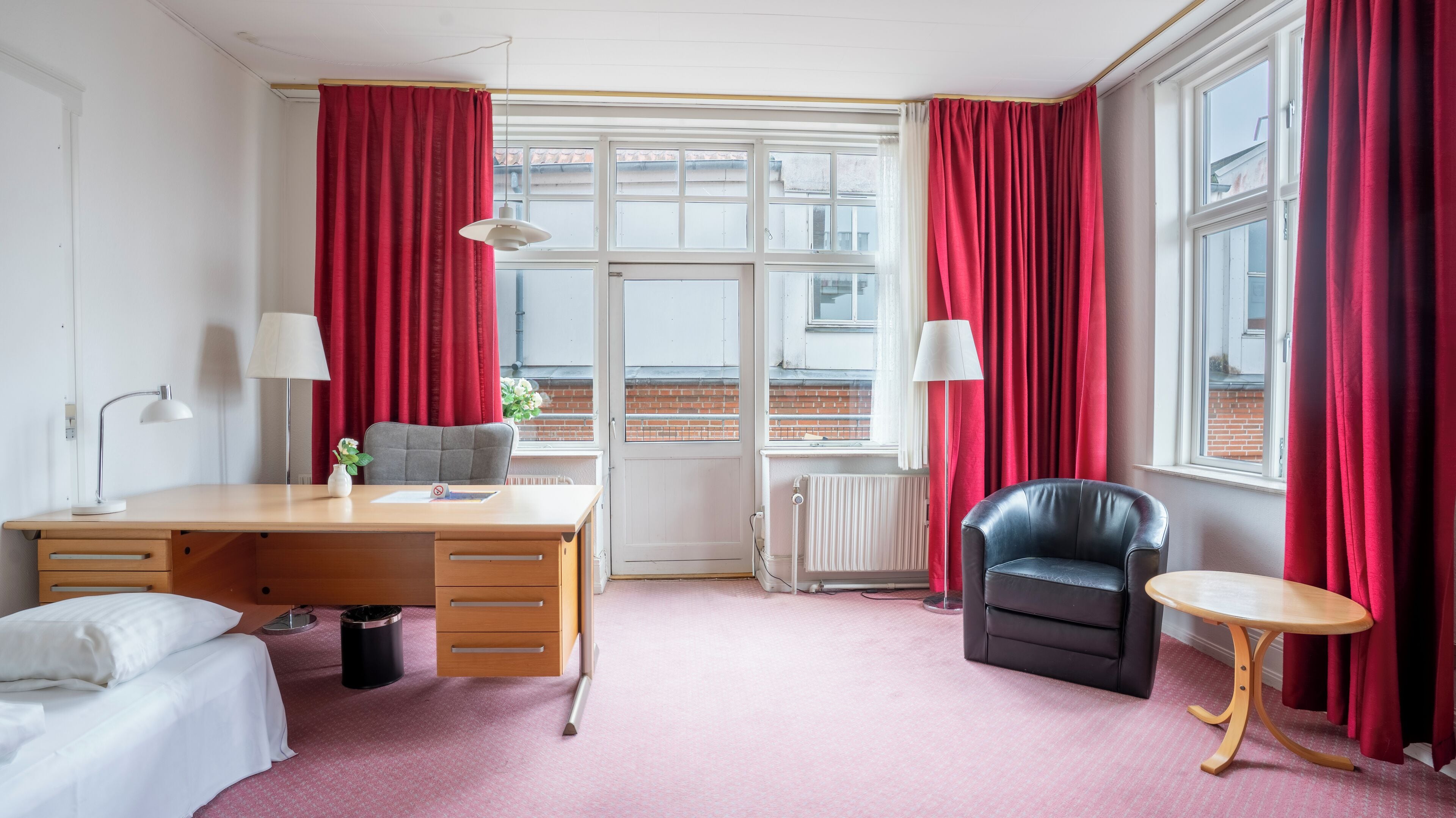 Une chambre d’hôtel avec un bureau en bois, deux chaises, une petite table, un radiateur et une fenêtre avec des rideaux rouges.