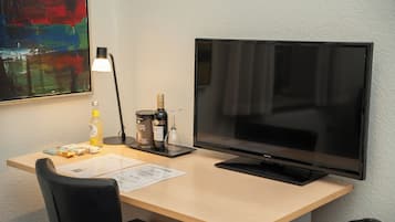 Ein Hotelzimmer mit Schreibtisch, Flachbildfernseher, Lampe, einer Weinflasche und einem Weinglas.