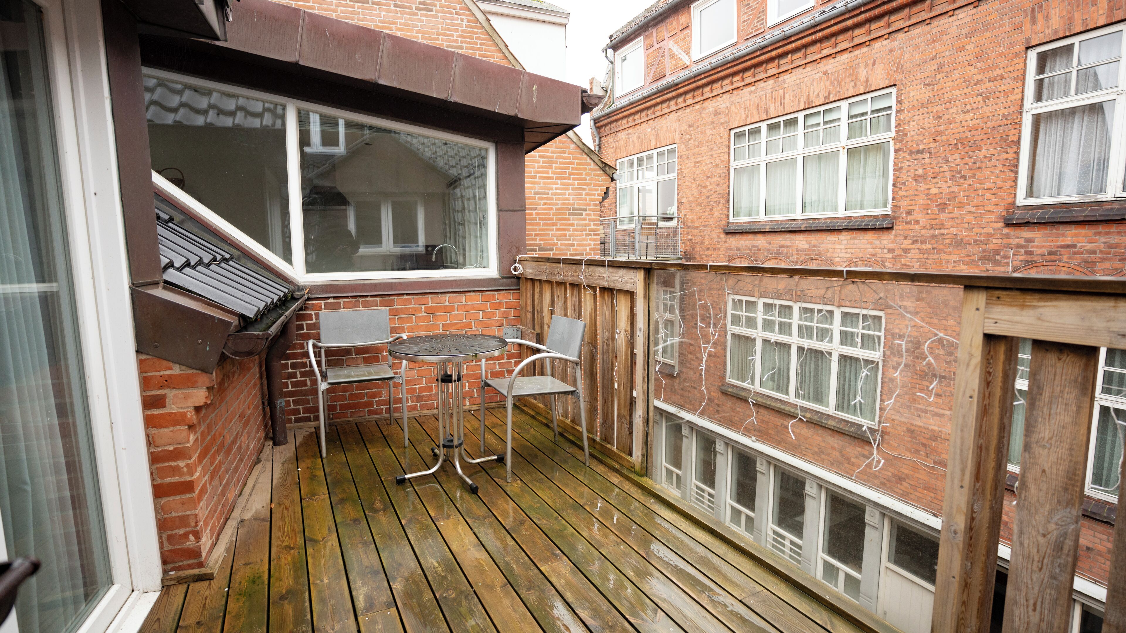 A wooden deck with two metal chairs and a small table, adjacent to a brick building with multiple windows.