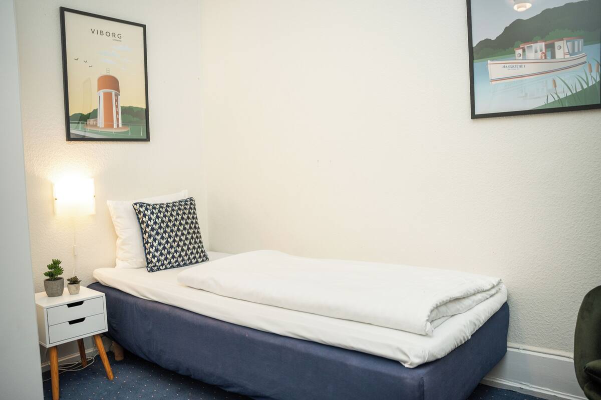 A neatly made bed with a white and blue bedspread, a patterned pillow, and a small bedside table with a lamp.