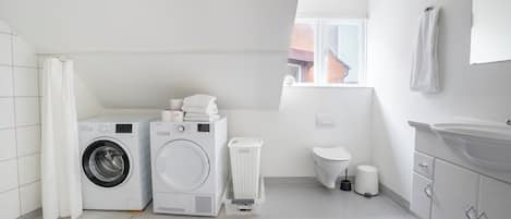 Een wasruimte met twee wasmachines, een droger, een witte wasmand, een toilet en een wastafel.