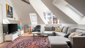 A modern loft apartment with a sloped ceiling, a large gray sectional sofa, a glass coffee table, and a colorful patterned rug. - Palads Hotel (Viborg)