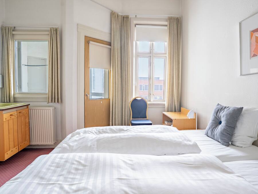 A hotel room with a bed, a desk, a chair, a radiator, and a window with curtains.