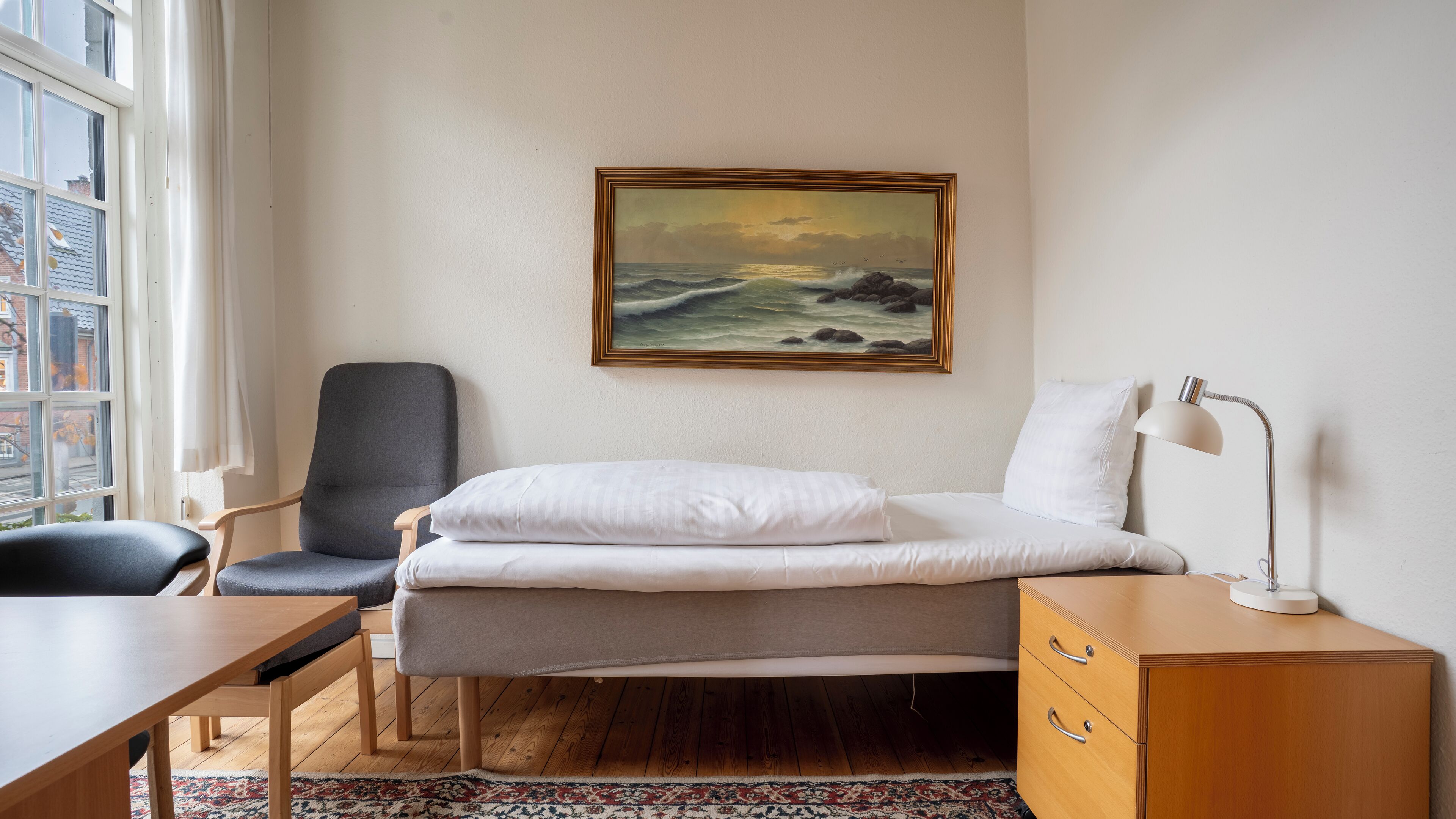 A room with a bed, a desk, a chair, a lamp, and a painting on the wall.