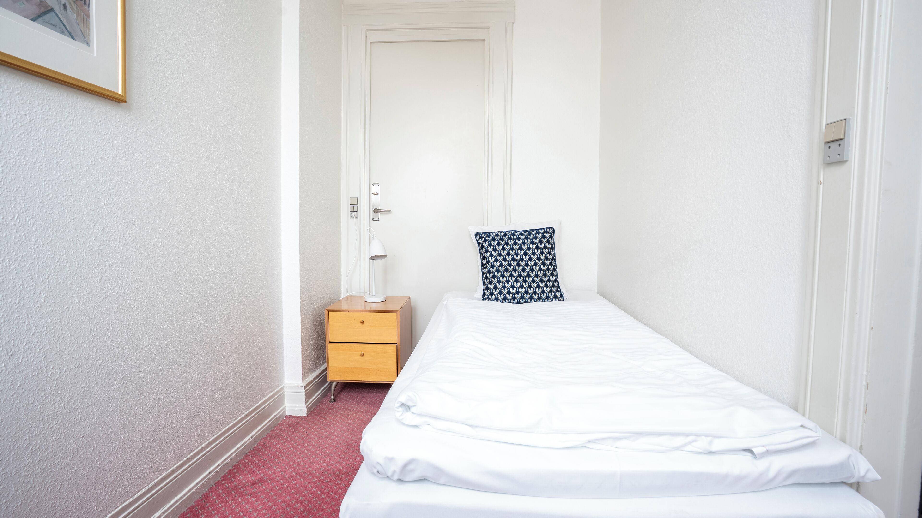 Une petite chambre d’hôtel étroite, avec un lit simple, une table de chevet, une lampe et une porte donnant sur une autre pièce.