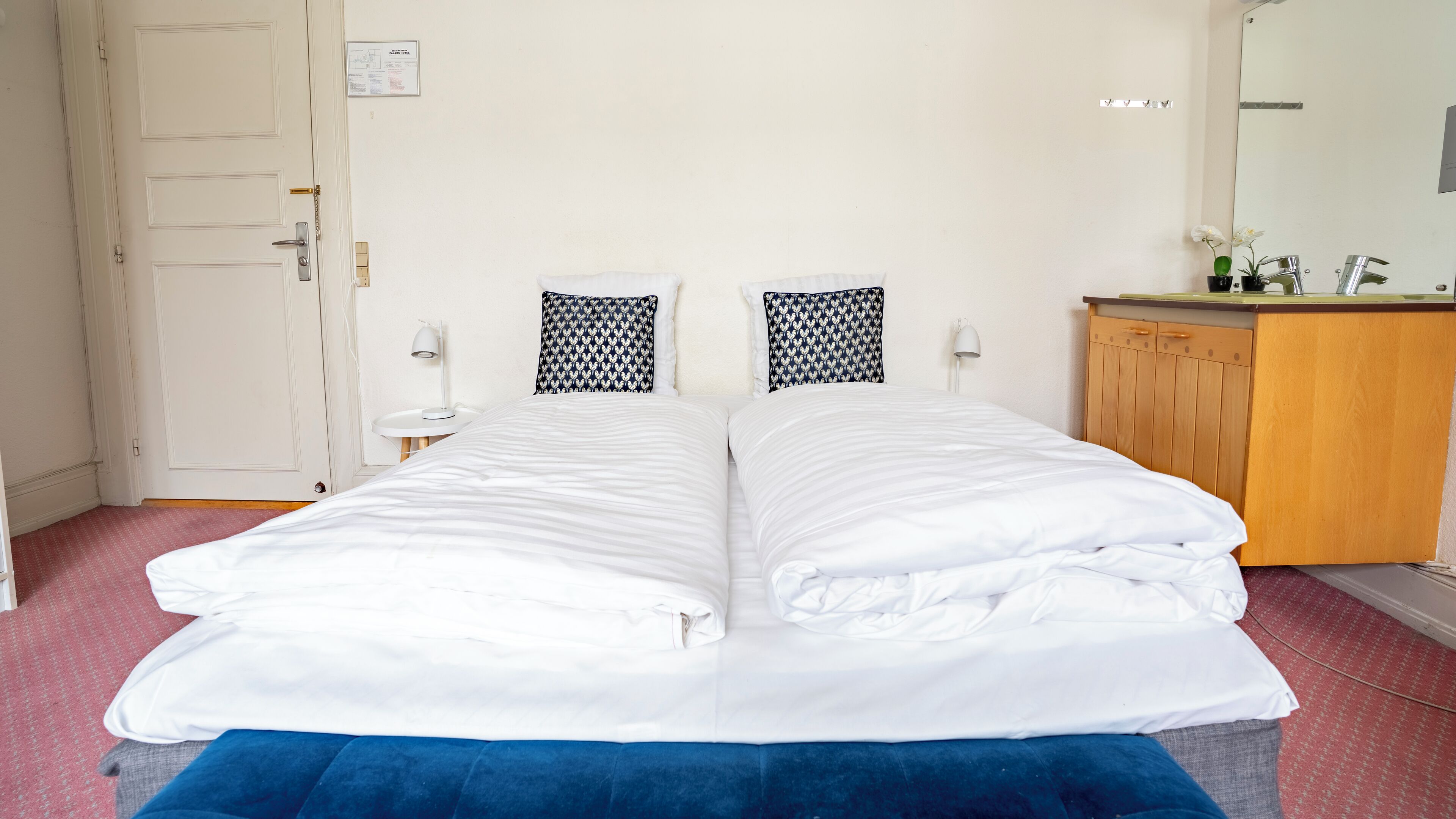 A double bed with white bedding, a blue accent pillow, a small bedside table with a lamp, and a wooden cabinet with a mirror.