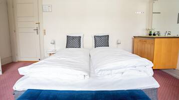 A double bed with white bedding, a blue accent pillow, a small bedside table with a lamp, and a wooden cabinet with a mirror.