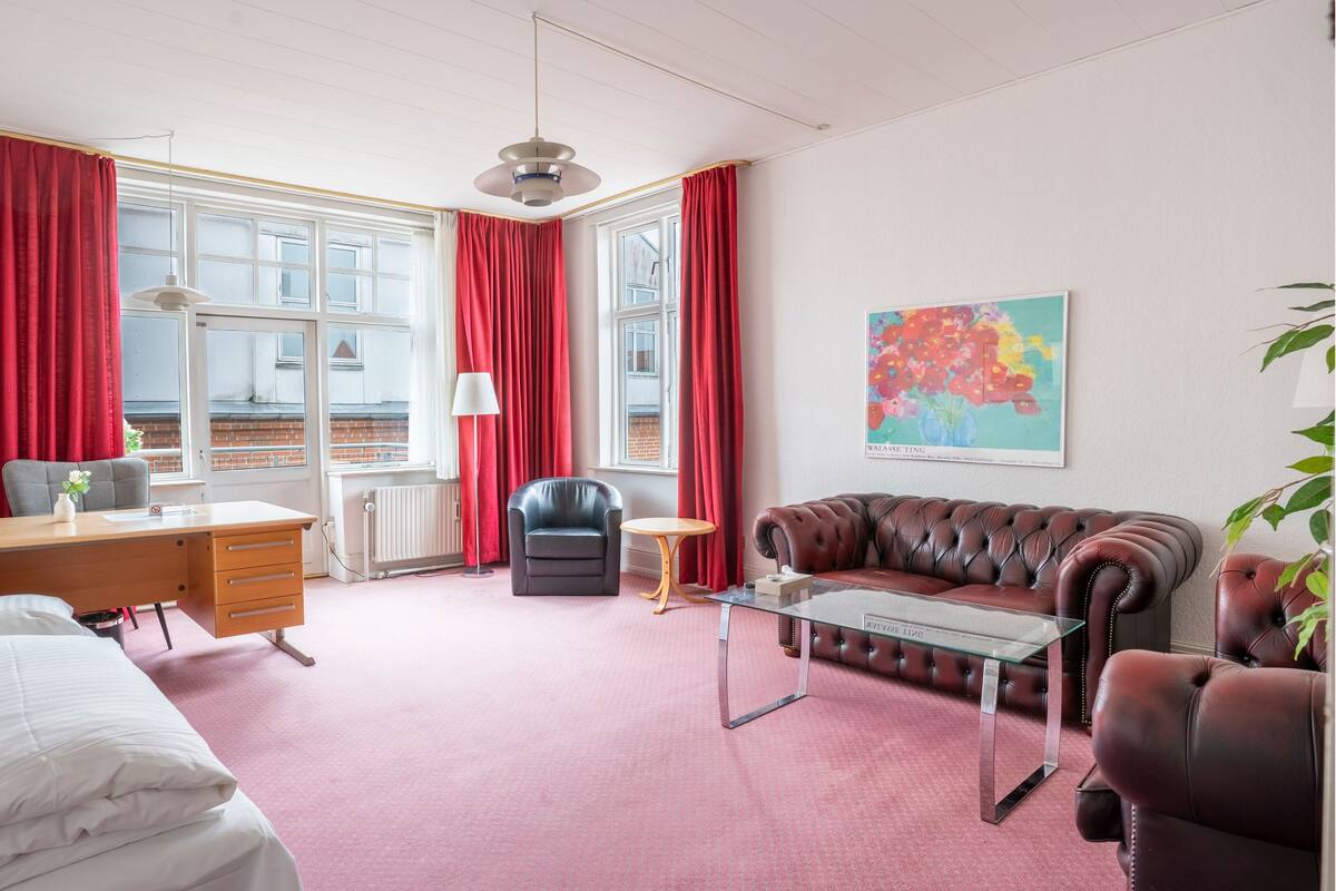 A hotel room with a bed, desk, chair, leather sofa, and a glass coffee table.