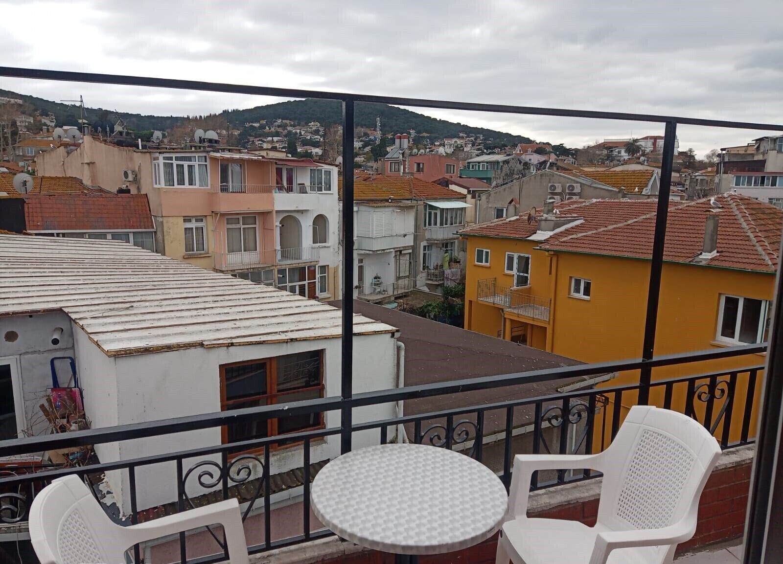 A balcony with a round table and two chairs, overlooking a residential area with colorful buildings and a hilly backdrop.