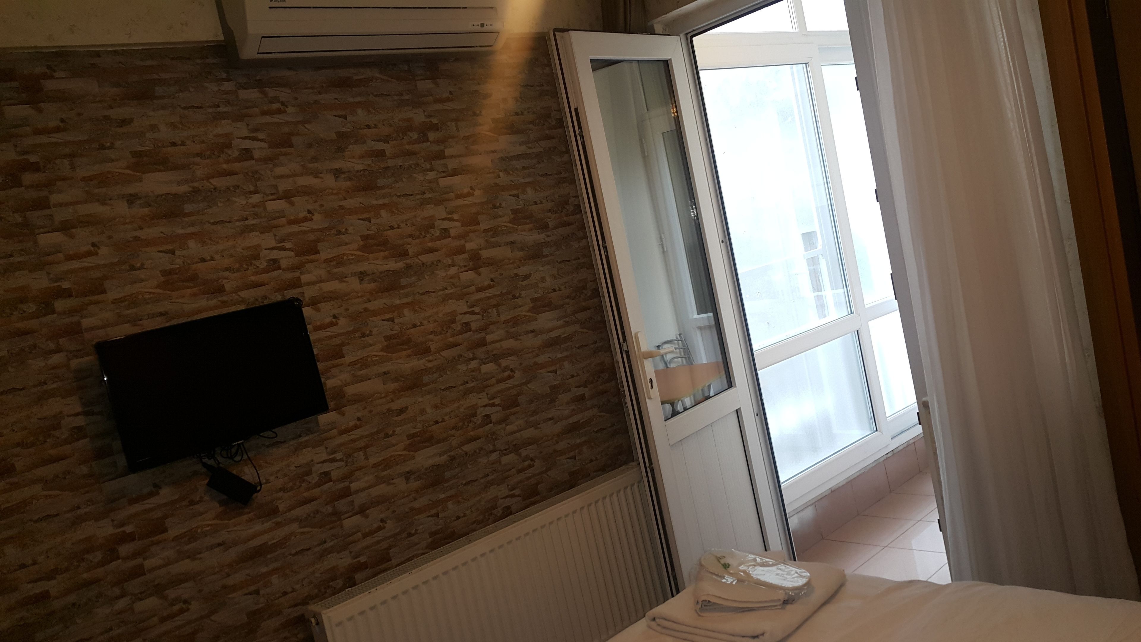 A hotel room with a brick wall, a flat-screen TV mounted on it, a radiator, a window with curtains, and a neatly made bed.