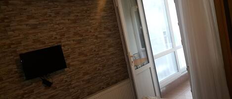 A hotel room with a brick wall, a flat-screen TV mounted on it, a radiator, a window with curtains, and a neatly made bed.