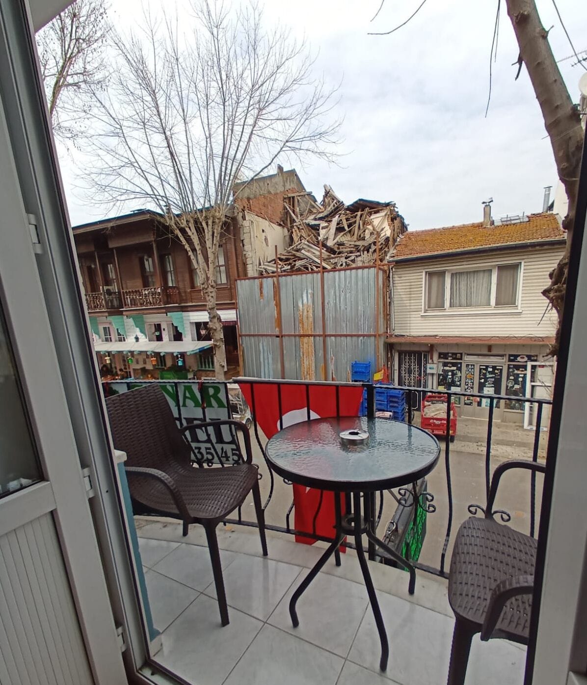 A balcony with a round table and two wicker chairs, a metal fence, and buildings in the background.