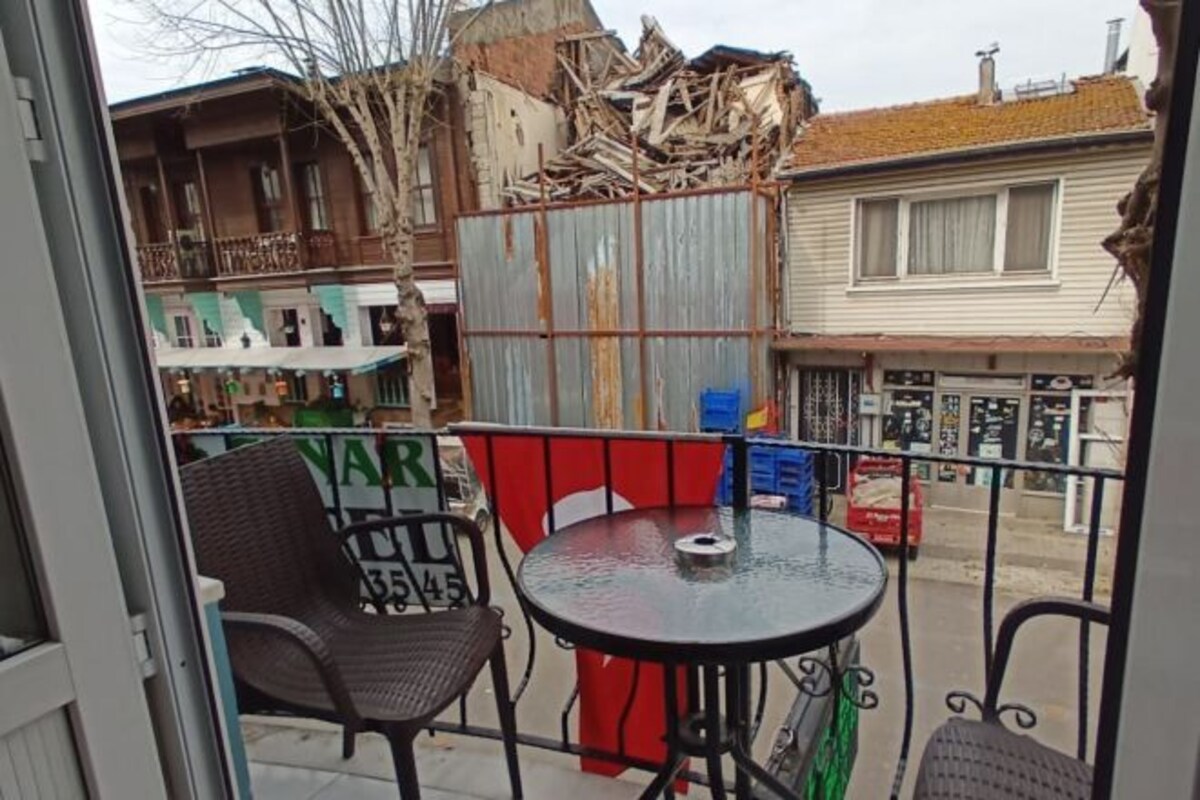 A balcony with a metal table and wicker chairs, overlooking a street with buildings and a pile of debris.