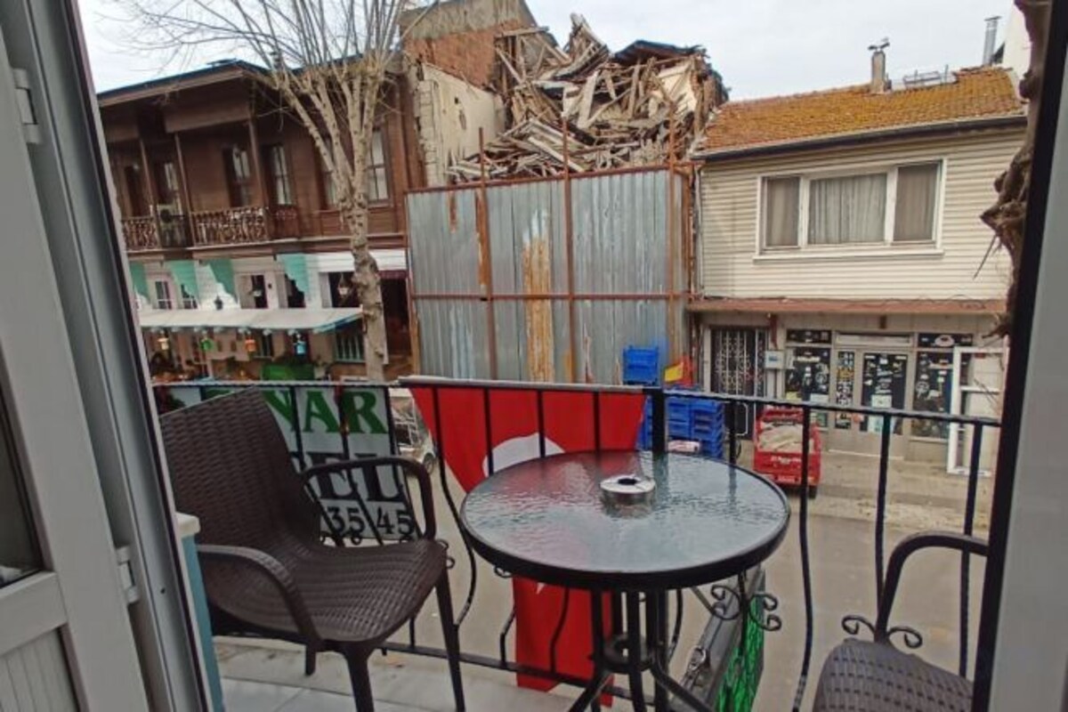 A balcony with a round table and two chairs, a metal railing, and a view of a building with a corrugated metal fence.