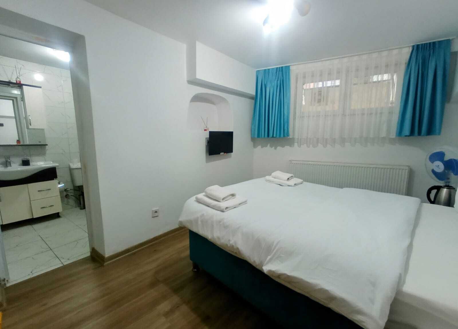 A bedroom with a bed, a small bedside table, a mirror, a window with curtains, a radiator, and a bathroom with a sink and mirror.