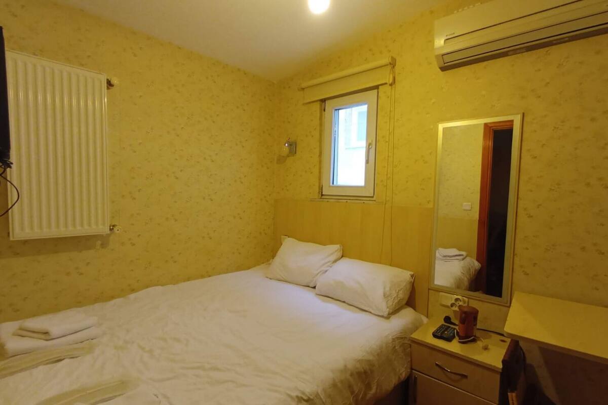 Une petite chambre avec un lit simple, une fenêtre, un miroir, un radiateur et une table de chevet sur laquelle se trouvent un téléphone et une tasse.