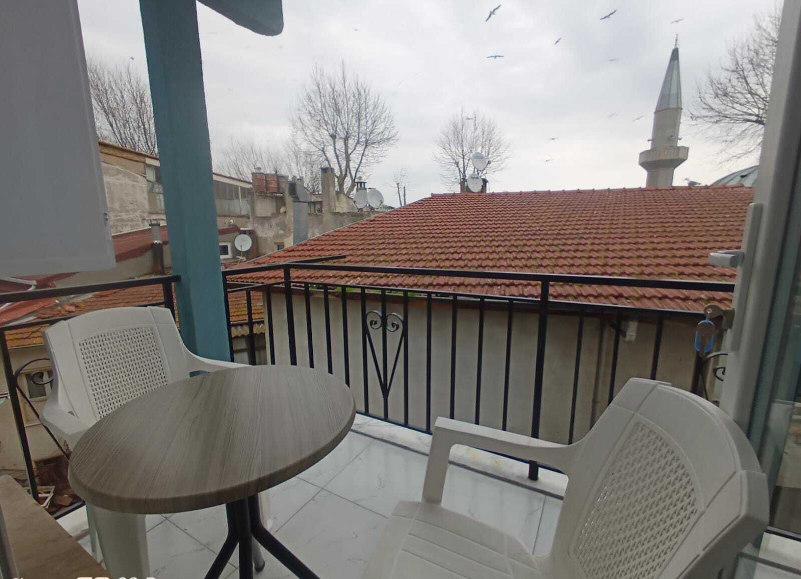 Un balcon avec une table ronde et deux chaises, d’où l’on peut admirer un bâtiment dont le toit est recouvert de tuiles rouges et qui est surmonté d’un minaret.