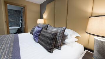 Habitación de hotel con cama, mesita de noche con lámpara, baño con bañera y puerta que da a otra habitación.