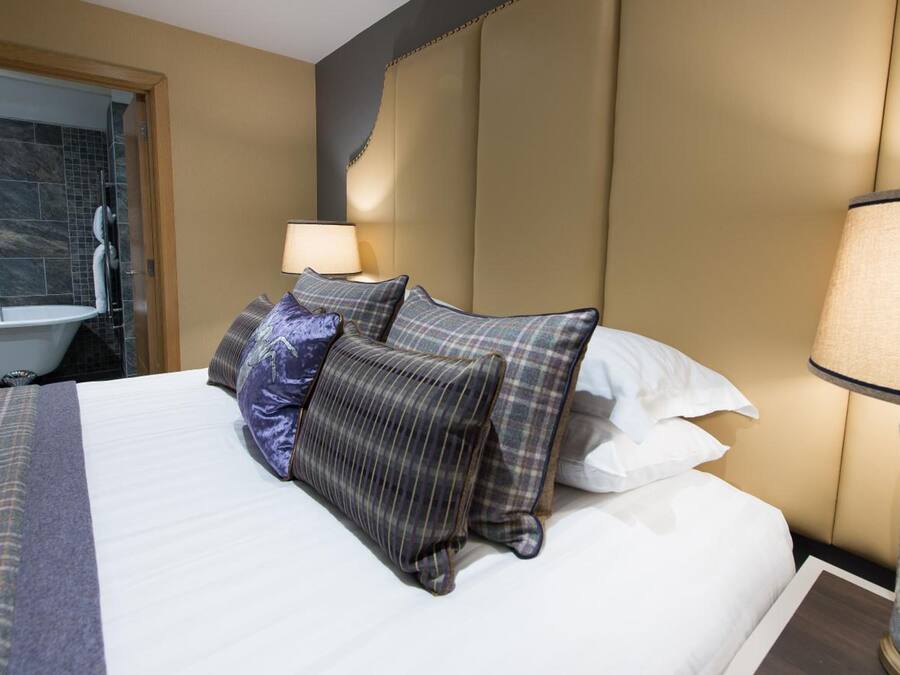 Habitación de hotel con cama, mesita de noche con lámpara, baño con bañera y puerta que da a otra habitación.