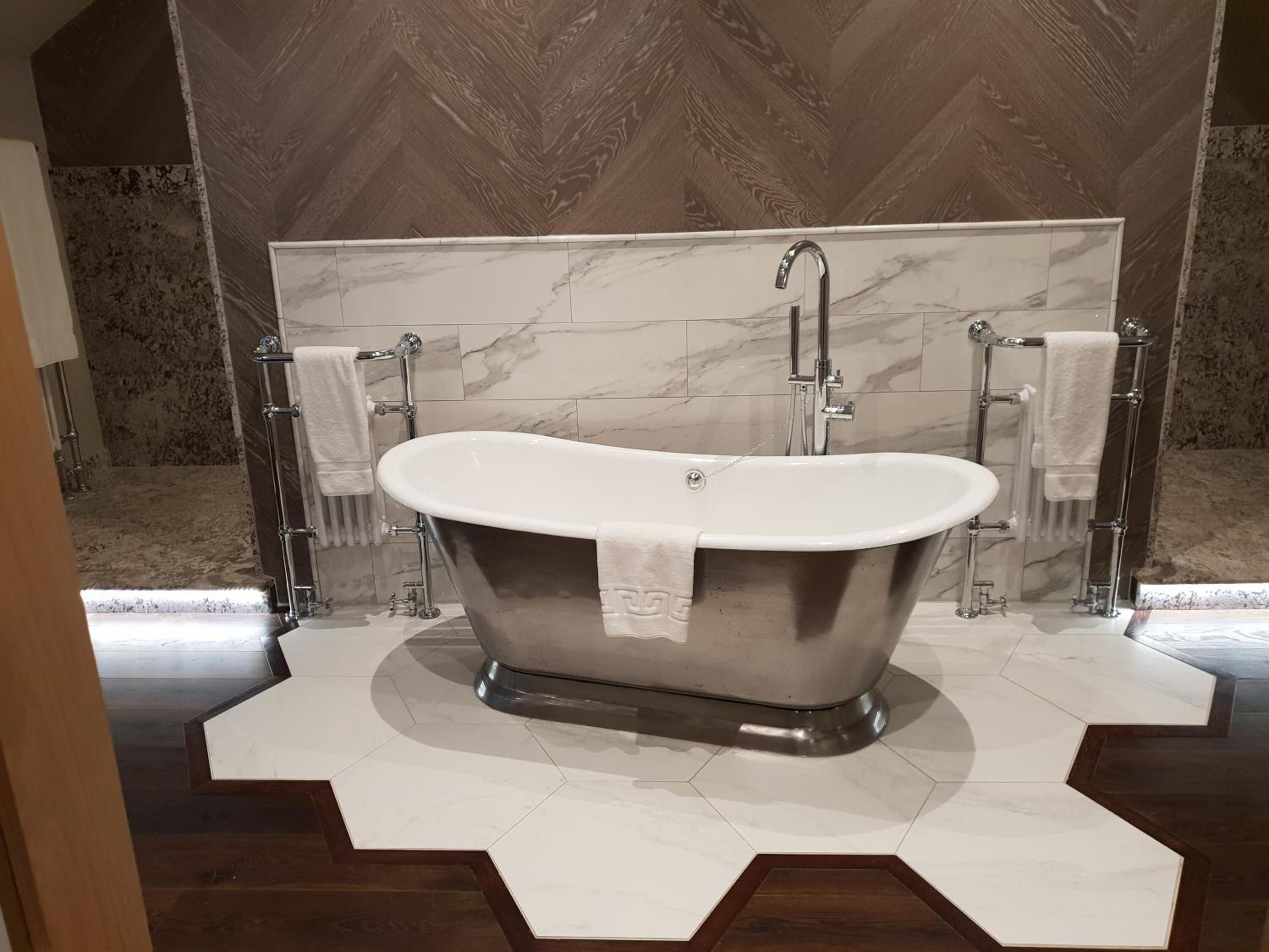 A freestanding bathtub in a modern bathroom with marble tiles and a herringbone-patterned wall.