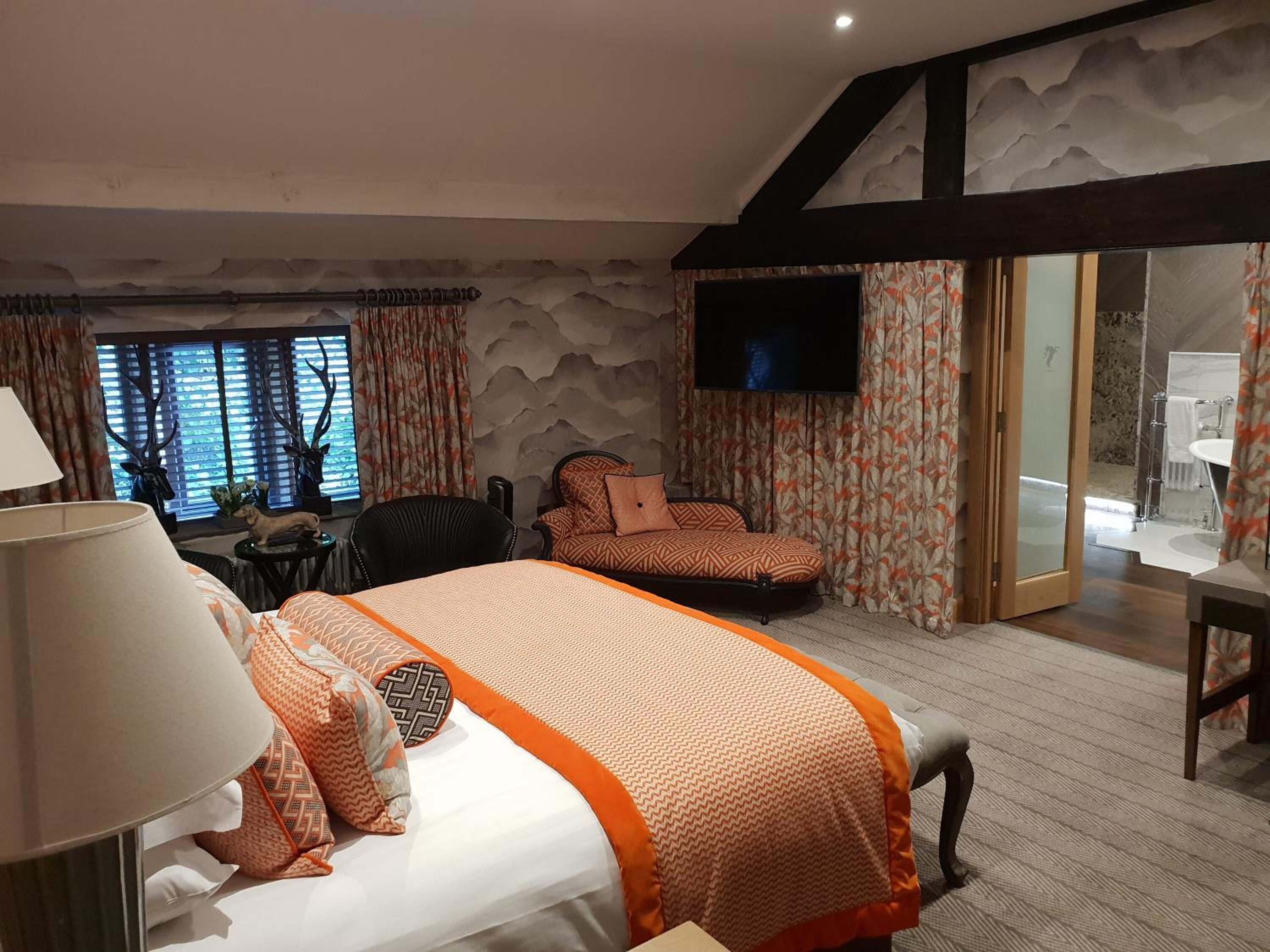 A hotel room with a bed, a desk, a chair, a TV, and a bathroom visible through a glass door.