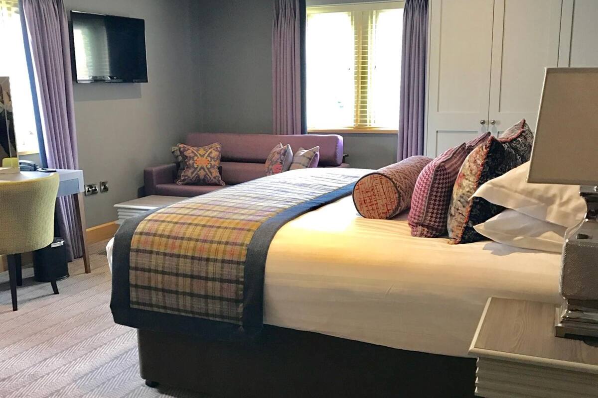 A hotel room with a large bed, a sofa, a TV mounted on the wall, and a desk with a lamp.