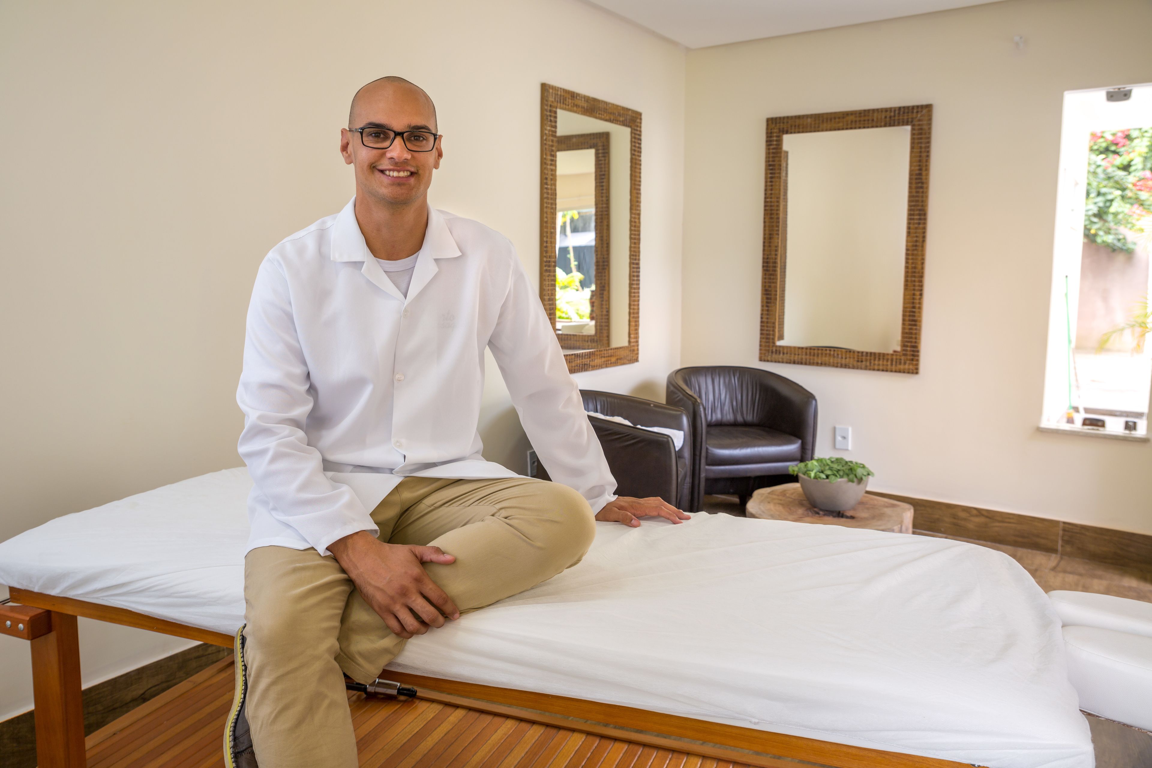 A man sitting on a bed in a room with a mirror, a chair, and a small table with a plant.