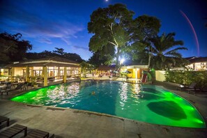 Une piscine éclairée de lumières vertes, entourée de chaises longues et d’un espace abrité avec des tables et des parasols.