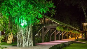 A covered walkway with a wooden deck and a green-lit tree. - Floral Inn Family Hotel (Porto Seguro)