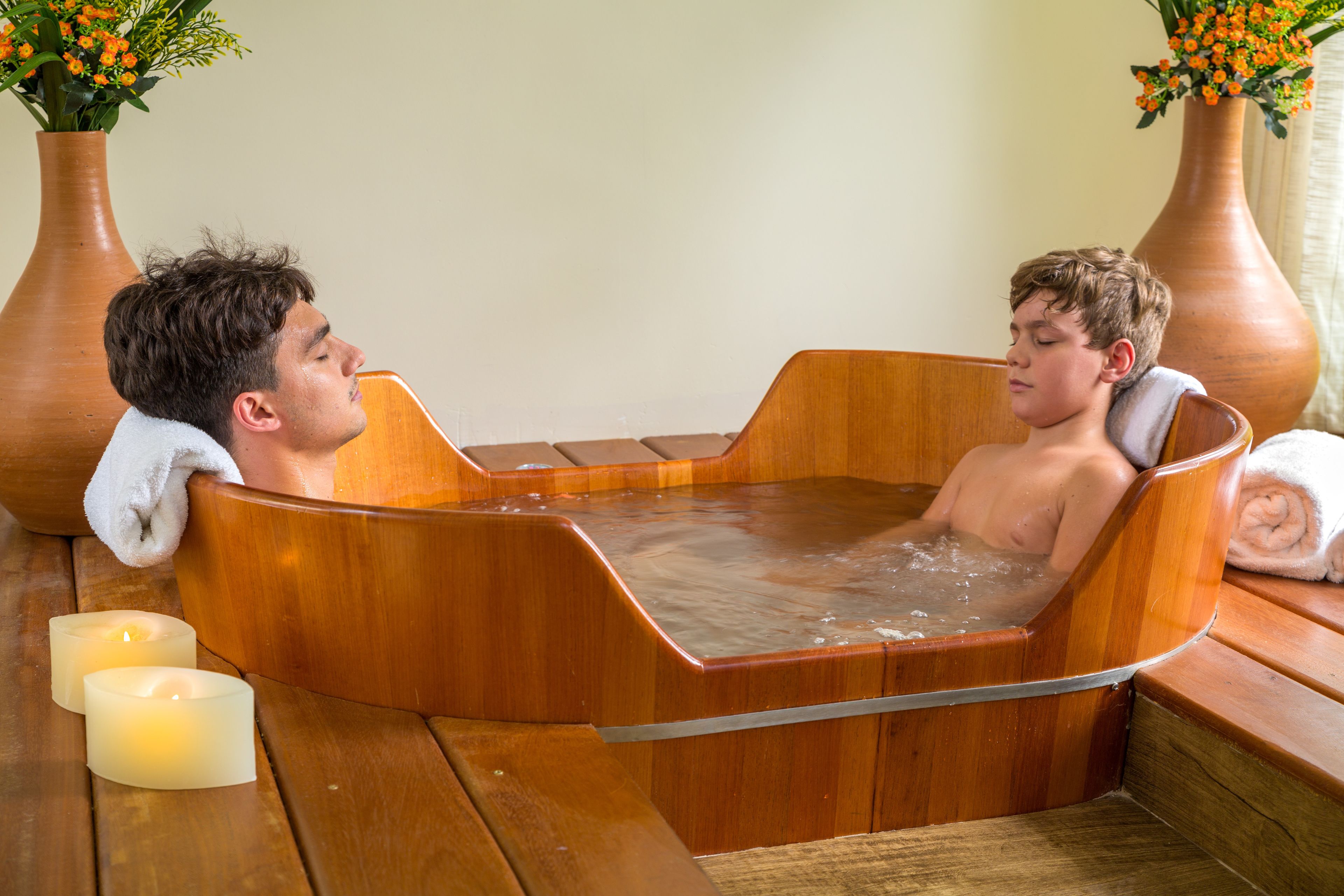 A wooden bathtub with two people inside, surrounded by candles and vases with flowers.