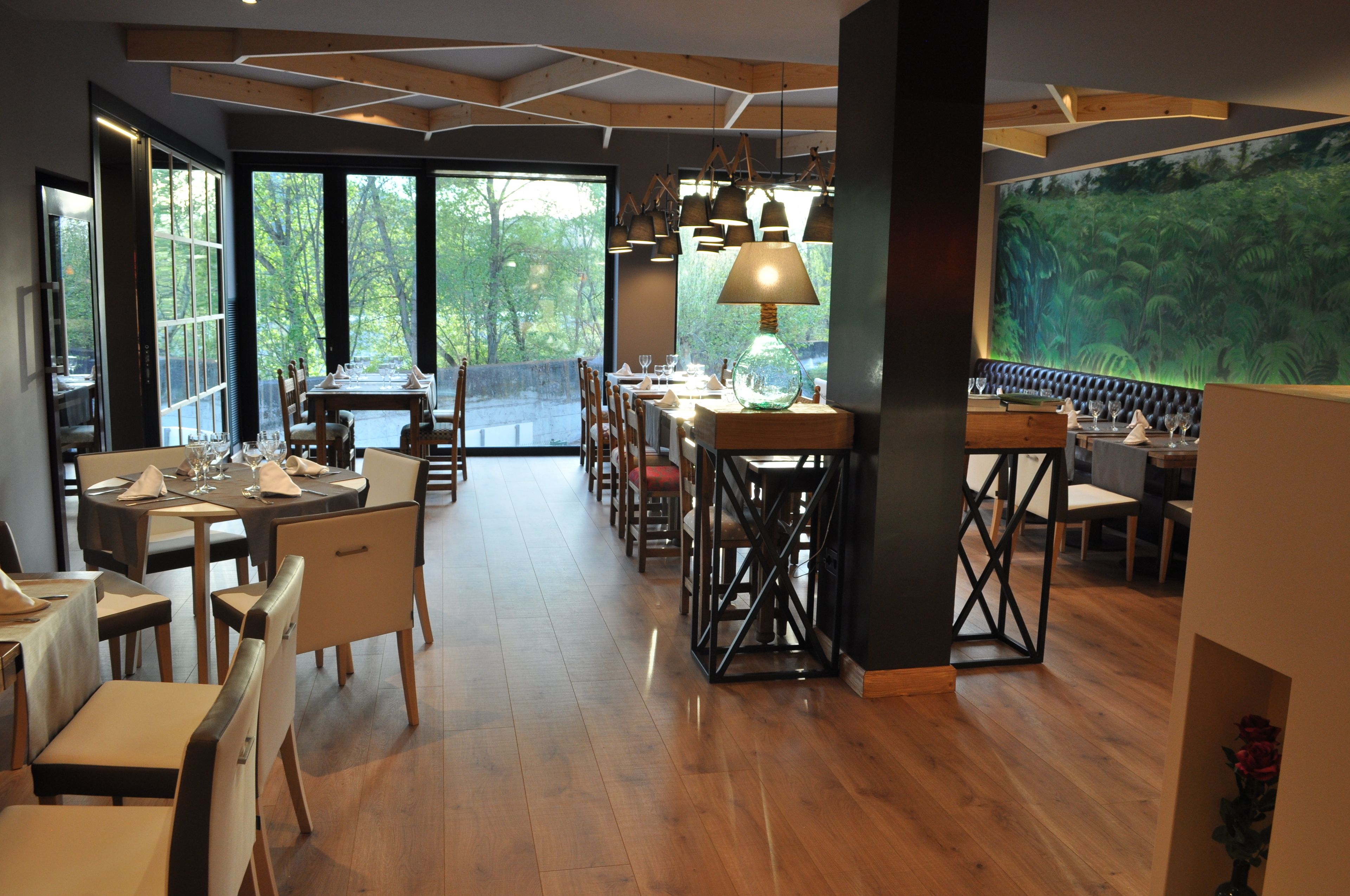 A restaurant with wooden flooring, large windows, and a view of greenery. Tables are set with white tablecloths and chairs.
