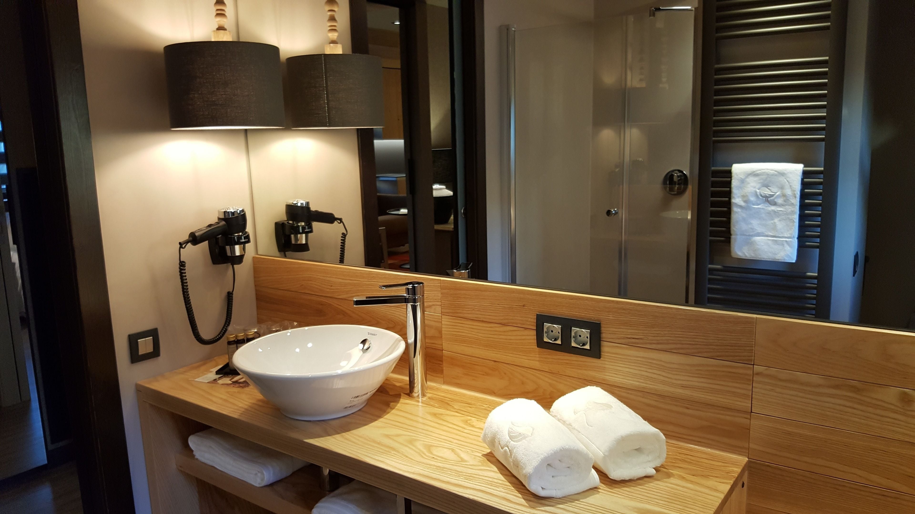 A modern bathroom with a wooden vanity, a white sink, and a mirror. There are two hair dryers on the wall, and a towel is rolled up on the vanity.