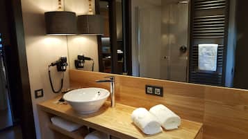 A modern bathroom with a wooden vanity, a white sink, and a mirror. There are two hair dryers on the wall, and a towel is rolled up on the vanity.