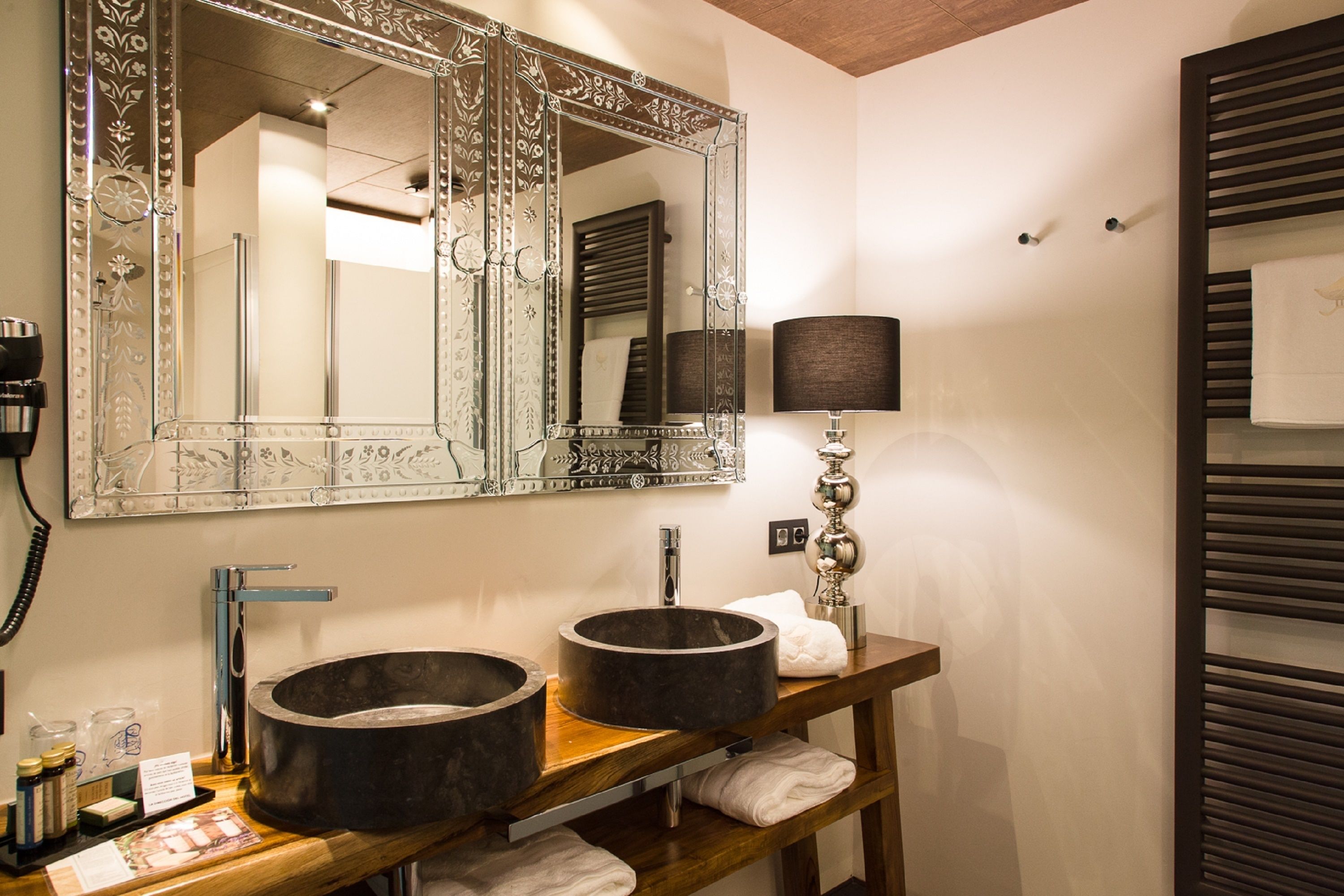A bathroom with two large, round sinks, a mirror with ornate framing, a towel rack, and a lamp.