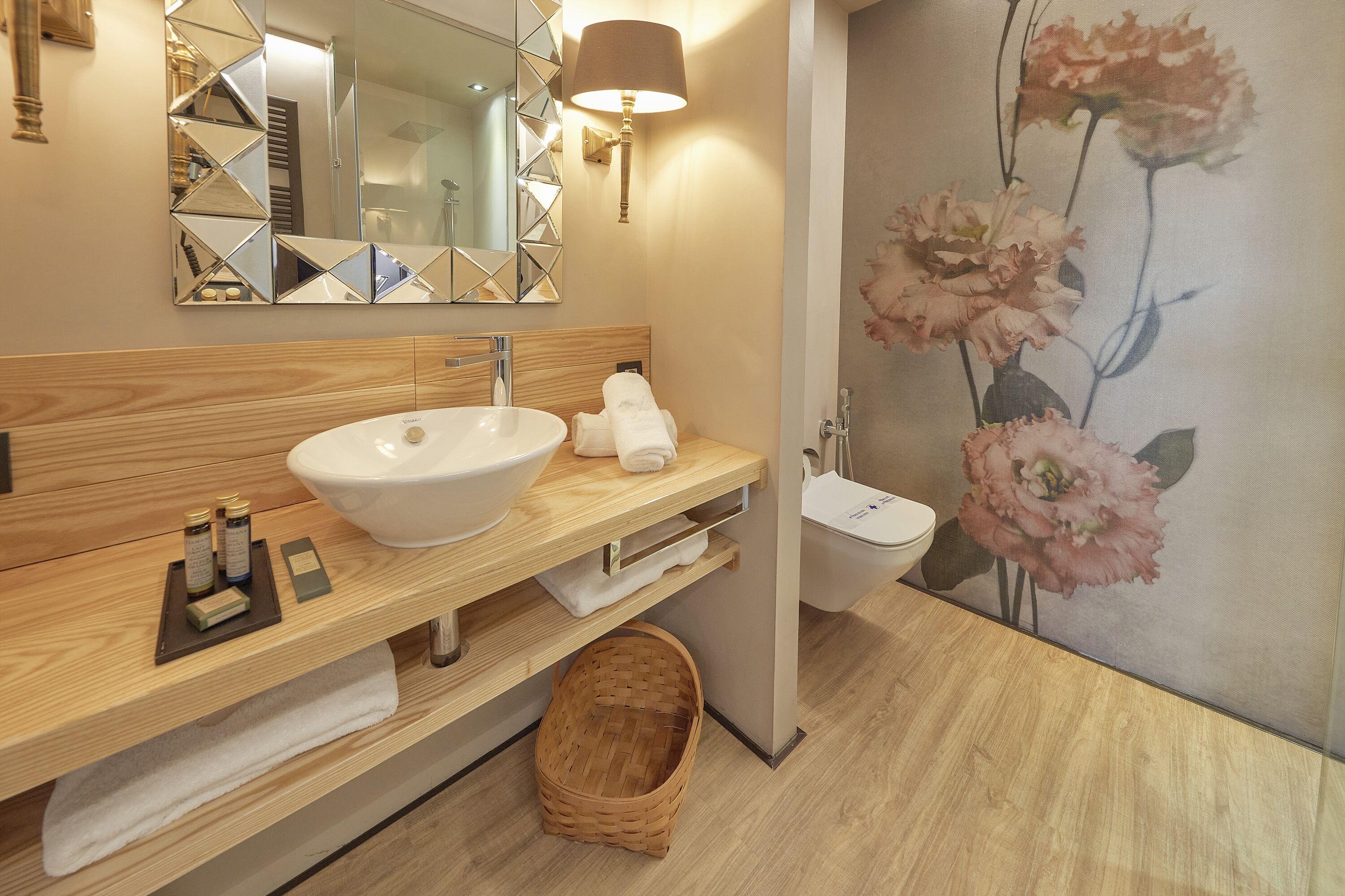 A modern bathroom with a wooden vanity, a white sink, a mirror with geometric patterns, a wall-mounted lamp, and a floral mural.