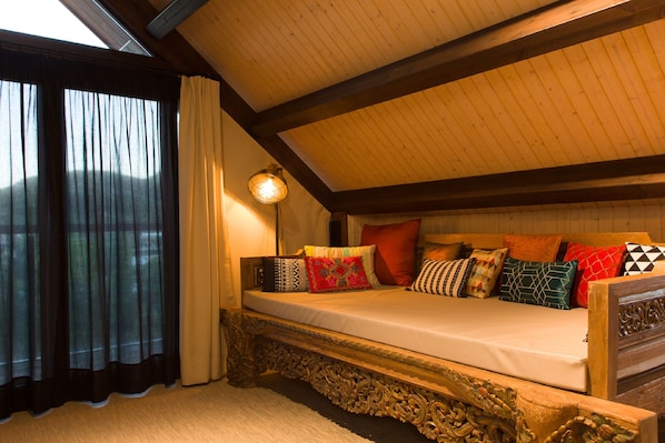 Un dormitorio acogedor con techo inclinado, una cama con muchos almohadones y una ventana con cortinas.