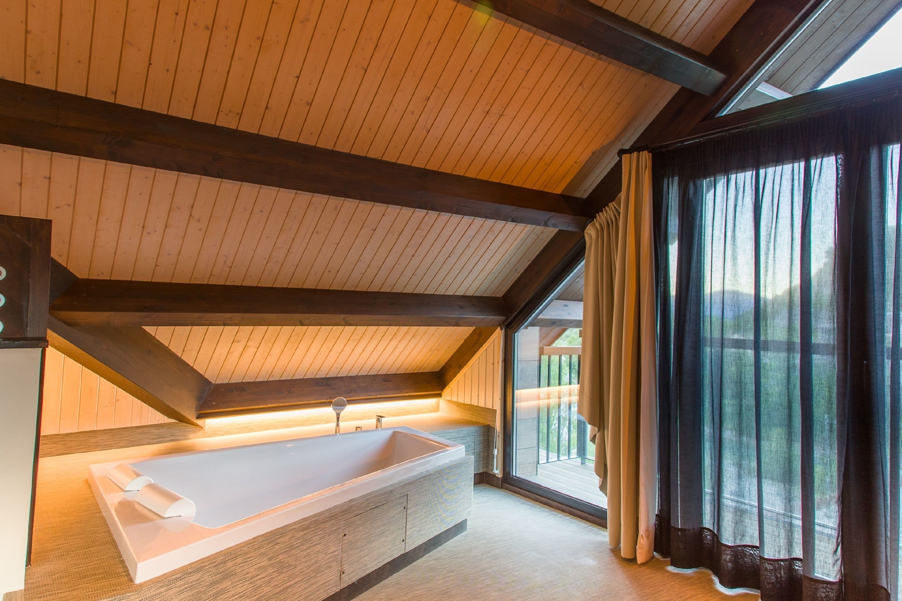 A modern bathroom with a freestanding bathtub, wooden ceiling beams, and a large window with curtains.