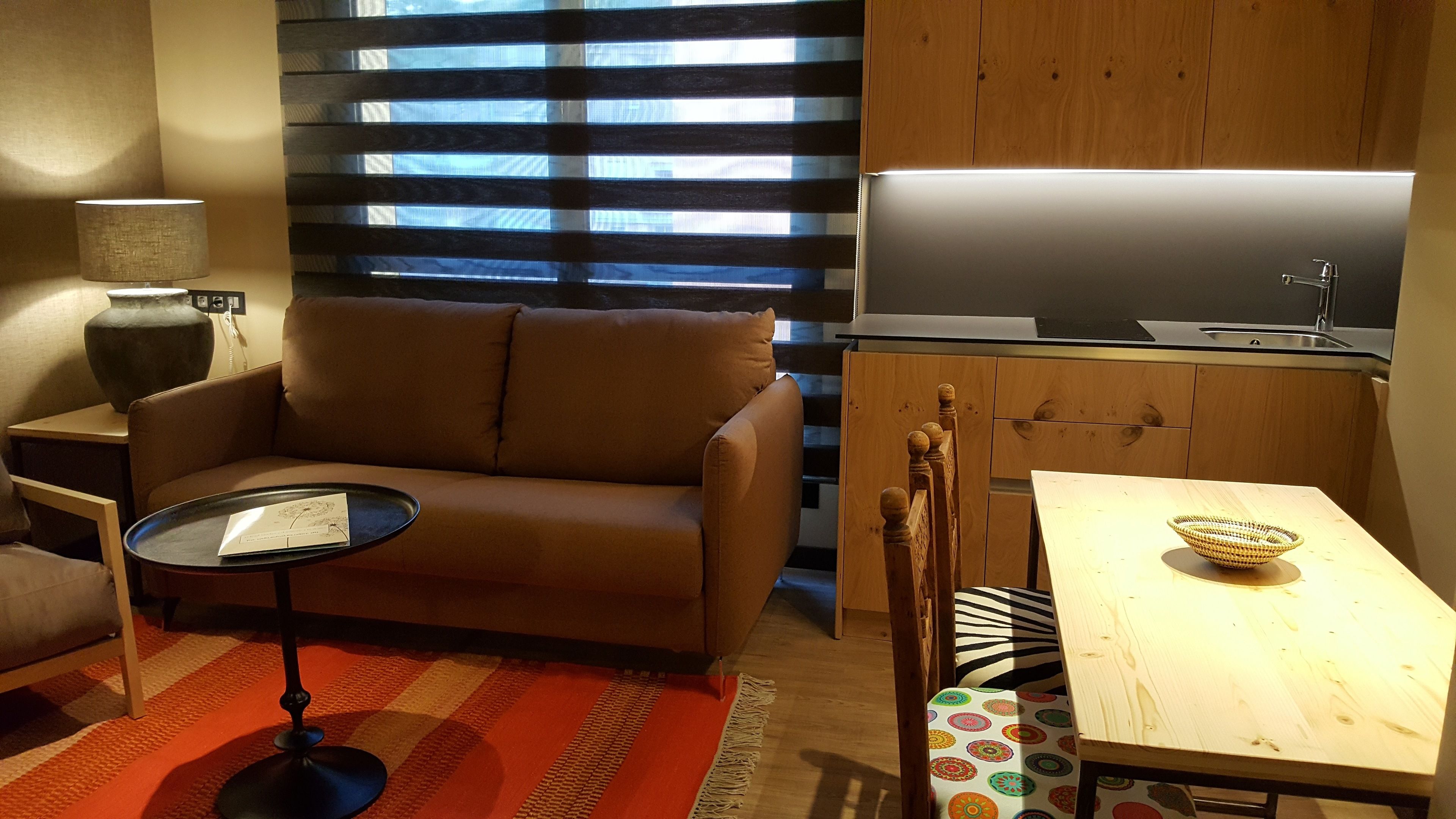 Un espacio habitable compacto con sofá, una mesita, una zona de cocina con fregadero y una ventana con persianas.