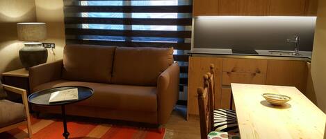 Un espacio habitable compacto con sofá, una mesita, una zona de cocina con fregadero y una ventana con persianas.