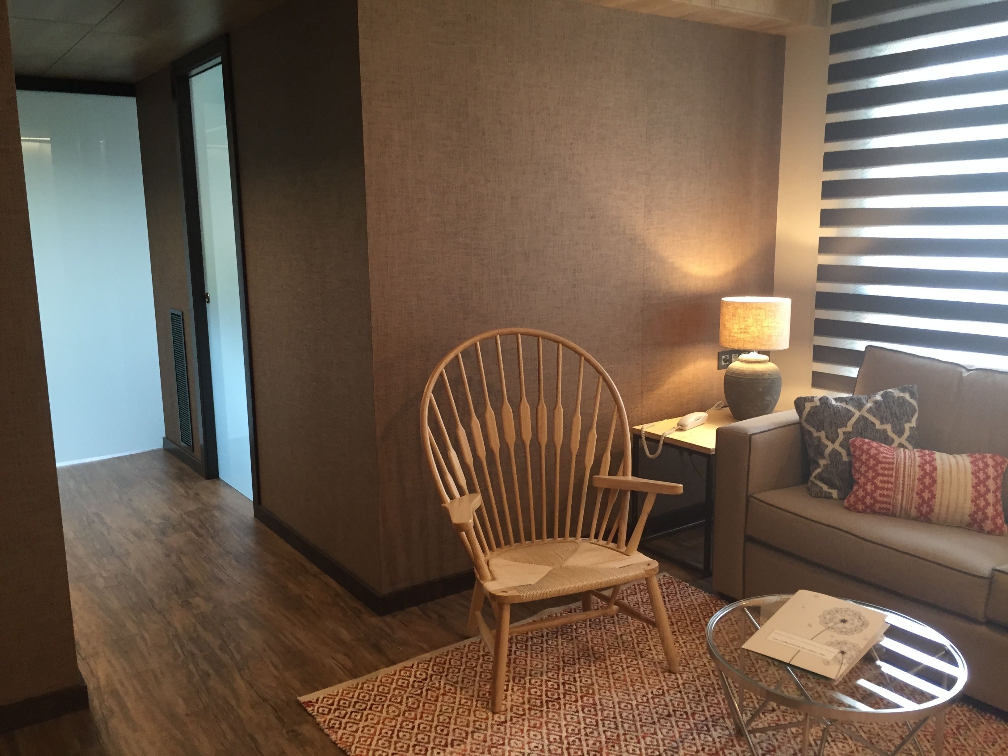 Una silla de madera, una mesa de vidrio con un libro, una lámpara y un sofá con almohadones estampados en una habitación con piso de madera y paneles en las paredes a rayas.