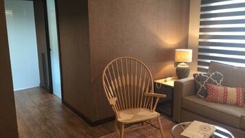 Una silla de madera, una mesa de vidrio con un libro, una lámpara y un sofá con almohadones estampados en una habitación con piso de madera y paneles en las paredes a rayas.